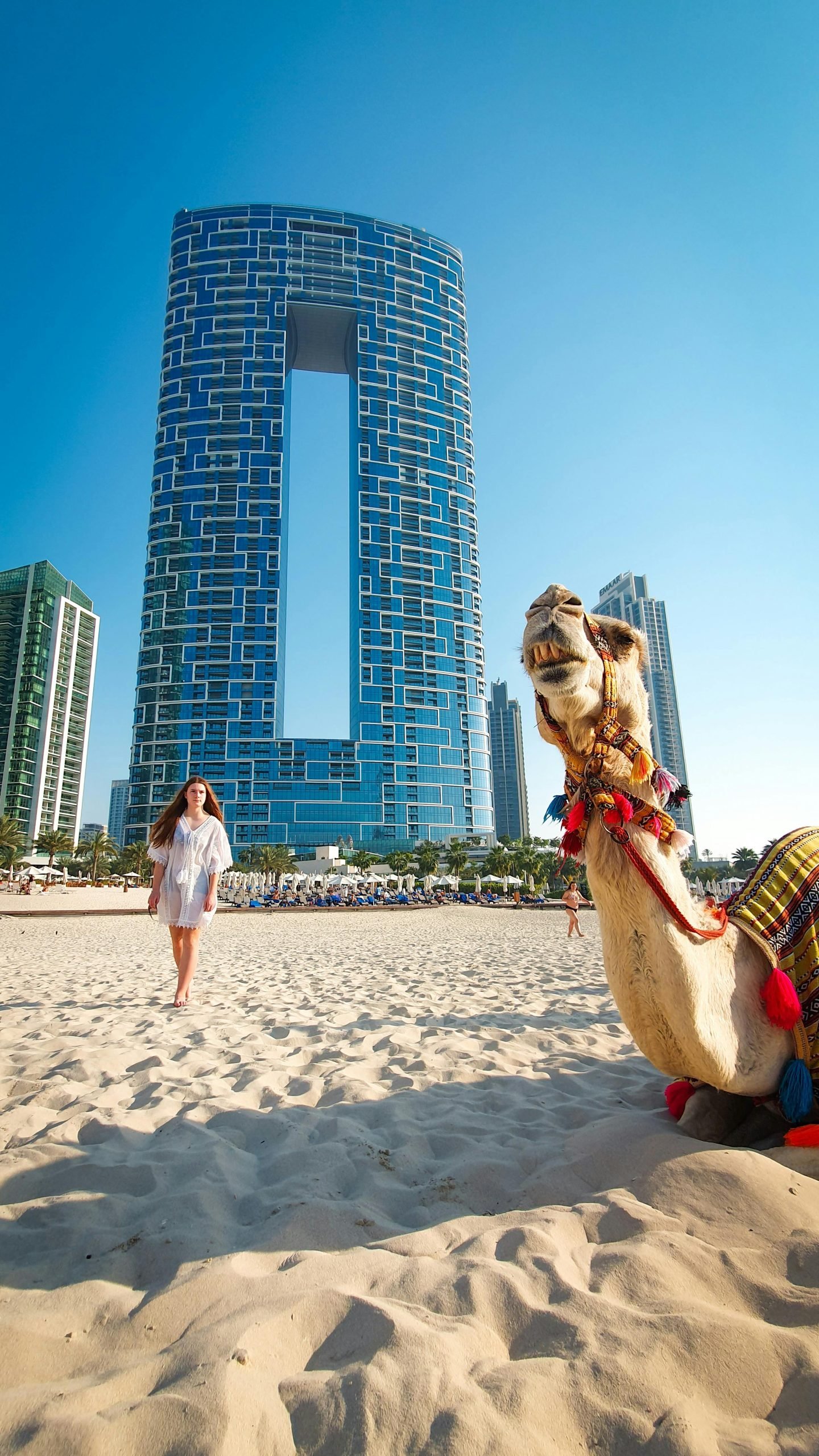 A woman walks on a Dubai beach with a camel and modern skyscrapers in the background.