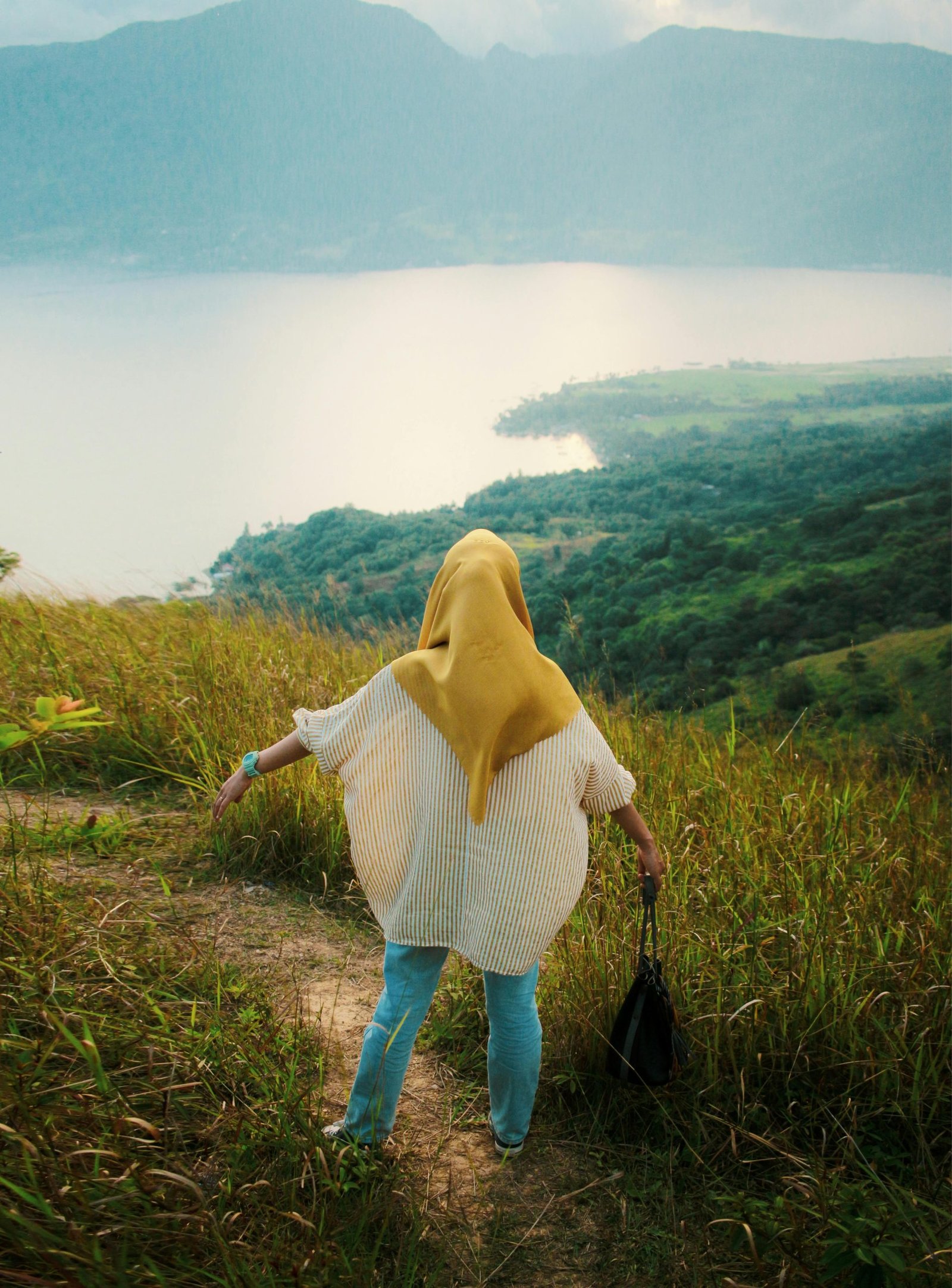 A woman in a hijab stands on a grassy hill, overlooking a serene lake and mountainous landscape at sunset.